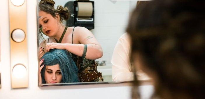 一位女演员在后台对着镜子帮另一位女演员戴假发