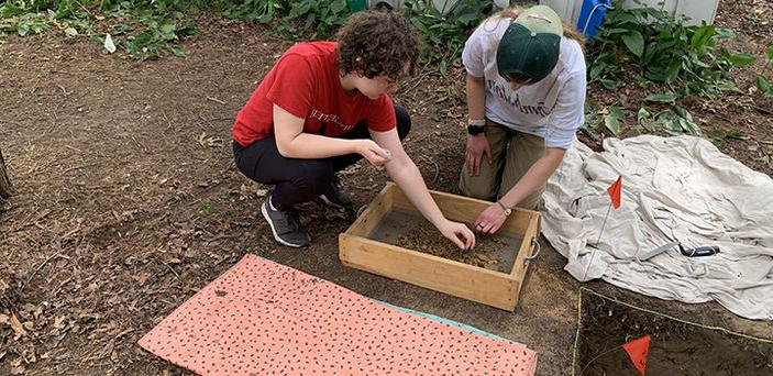 两名波士顿州立大学考古学专业的学生跪在一块泥土上，用从他们在地上挖的洞里取出的筛子拣东西